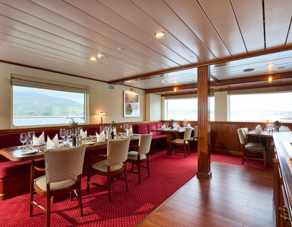 Restaurant mit Holzvertäfelung, rotem Teppich, gedeckten Tischen und Fensterblick auf eine bergige Küstenlandschaft