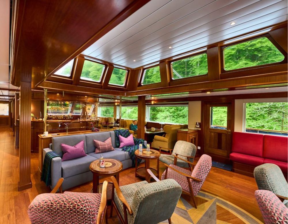 Lounge mit Mahagoni-Vertäfelung, Sofa, gemusterten Sesseln und Bar sowie Panoramafenstern mit Blick auf bewaldete Ufer