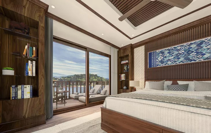 Geräumige Kabine mit Holzvertäfelung, Doppelbett und Bücherregal auf einem Flusskreuzfahrtschiff, dahinter ein privater Balkon mit Liegen und Blick auf den Mekong.