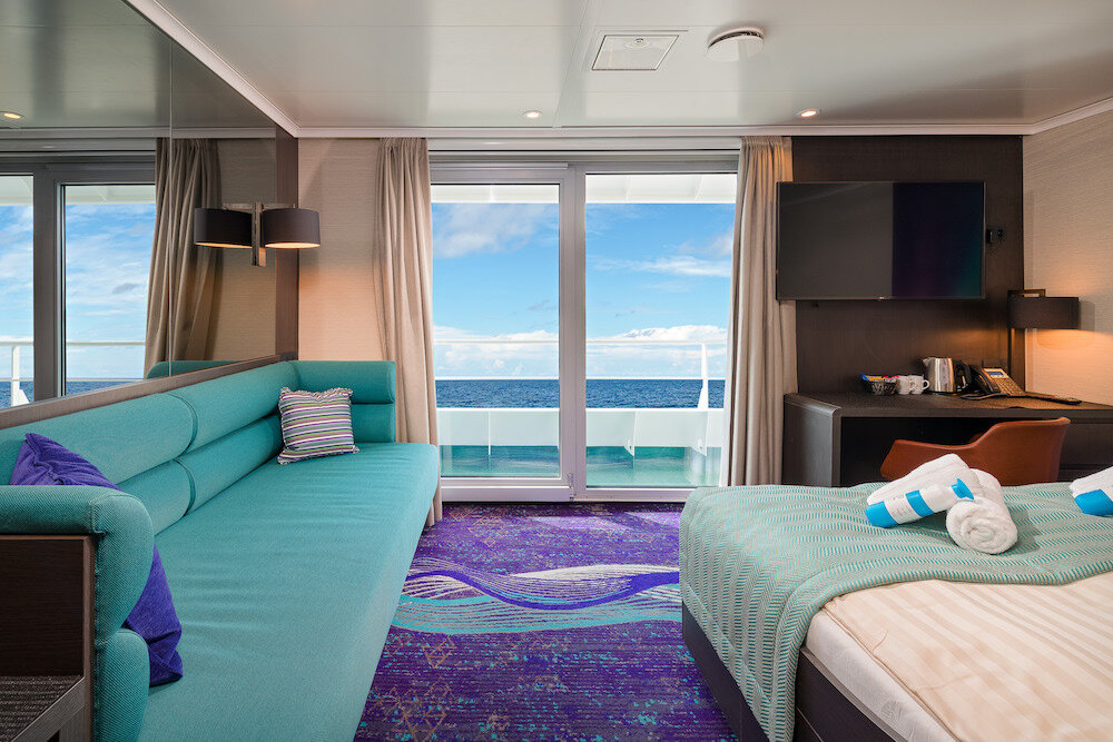 Suite mit Sofa, Doppelbett und Schreibtisch sowie Schiebetür zum eigenen Balkon mit Blick auf das offene Meer.