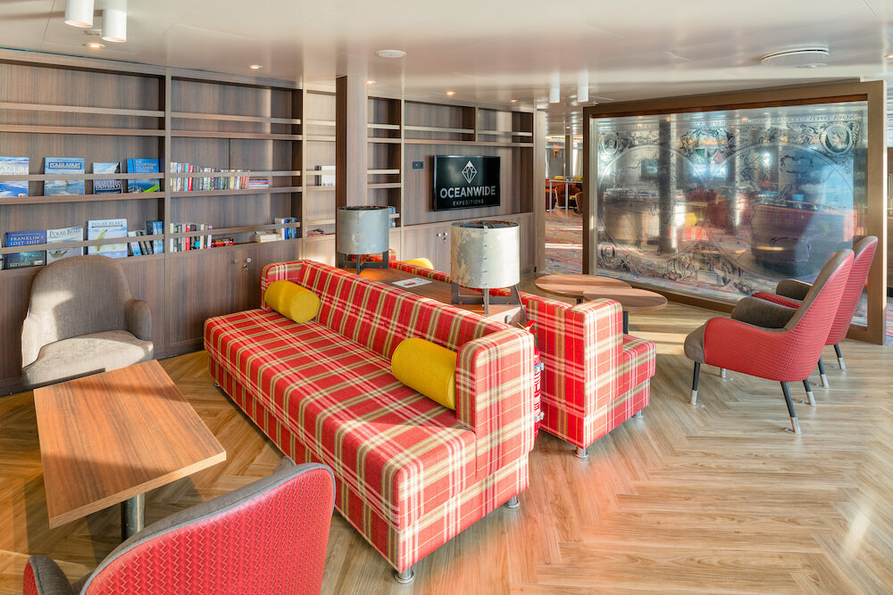 Bibliothek und Loungebereich an Bord eines Expeditionsschiffs mit Bücherregalen, kariertem Sofa und Sesseln.