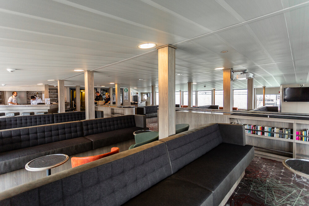 Aussichtslounge eines Expeditionsschiffs mit langen Sitzbankreihen, Bücherregal, Beamer und Panoramafenstern, zugleich Vortragsraum.