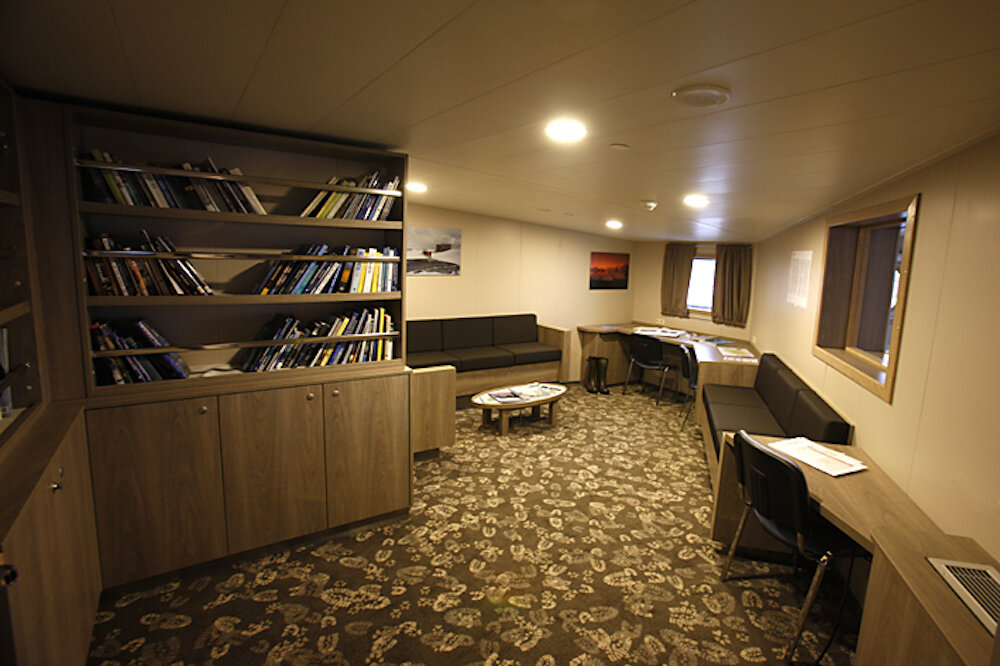Bibliothek an Bord mit großem Bücherregal, gepolsterten Sitzbänken, Couchtisch und Schreibtischen entlang der Wand.