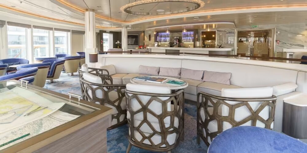 Helle Beobachtungslounge mit Sofa, Sesseln, Seekarten und Bar im Hintergrund mit Panoramafenstern