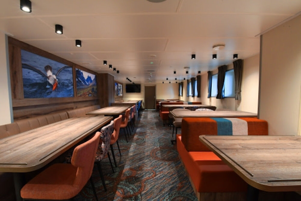 Speisesaal eines Expeditionsschiffs mit langen Holztischen, Sitzbänken und Bildschirm für Vorträge.