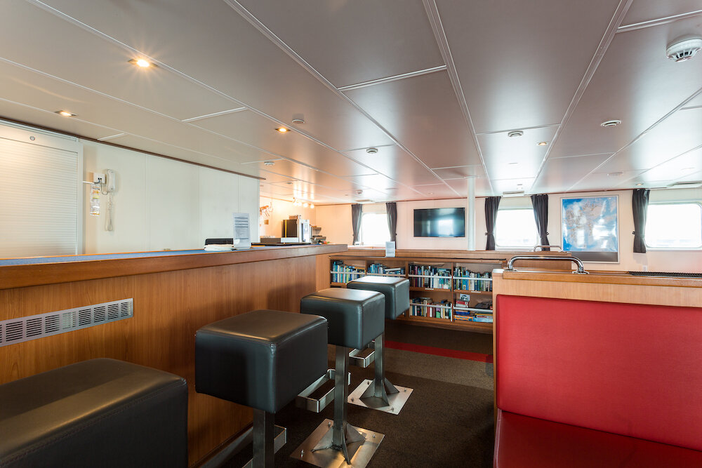 Aussichtslounge eines Expeditionsschiffs mit Bartresen, Barhockern, Bücherregal, Sitzbänken und Fenstern zur Seeseite.