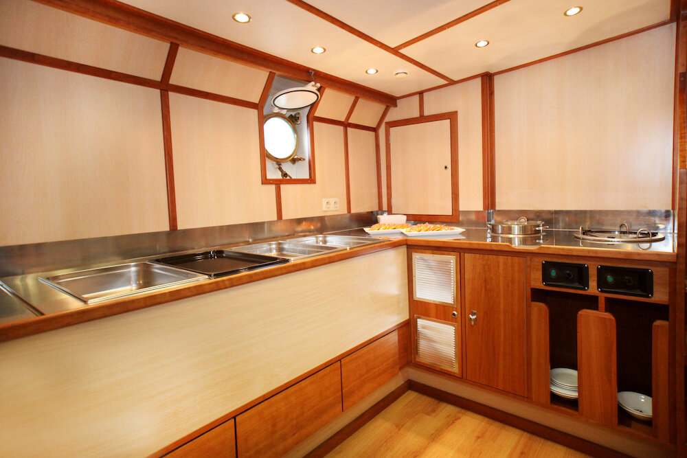 Buffetbereich mit Edelstahltheke, Warmhaltebehältern und Bullauge an Bord eines Segelschiffs.