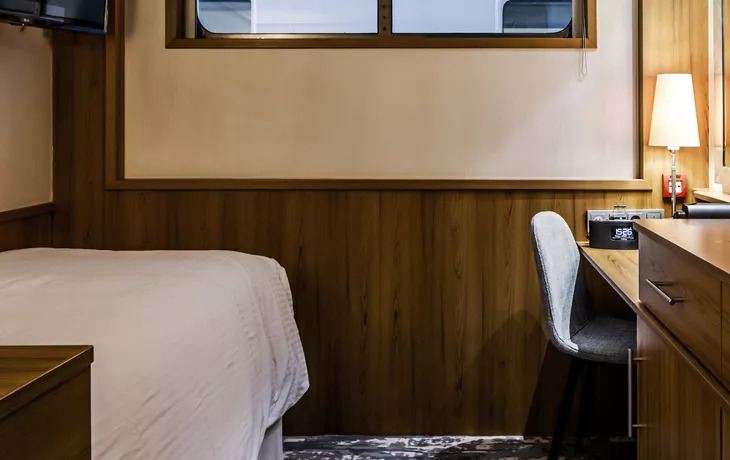 Einzelkabine auf dem Hauptdeck mit Bett, Schreibtisch mit Stuhl und schmalem Fenster über Holzvertäfelung
