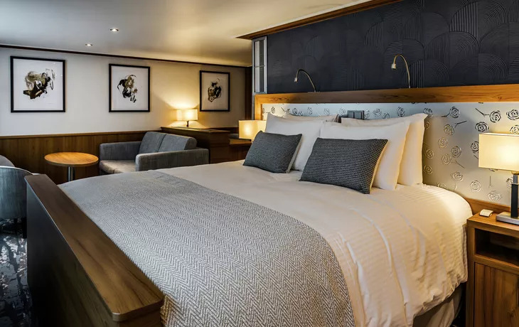 Geräumige Suite eines Flusskreuzfahrtschiffs mit Doppelbett, Sitzecke mit Sessel und Beistelltisch sowie Holzvertäfelung und Fensterfront.