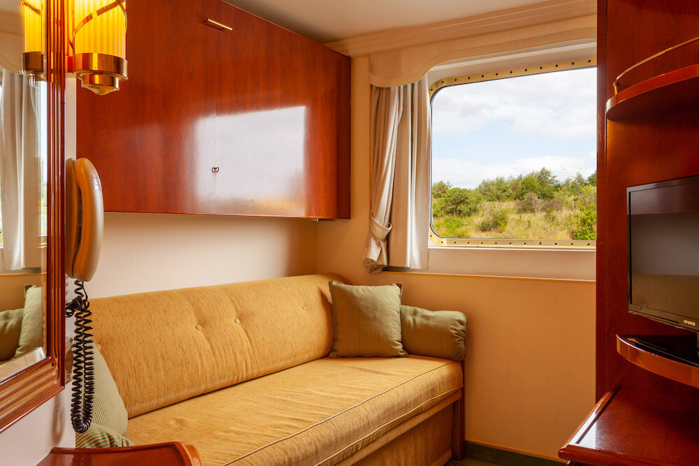 Einzelkabine auf dem Hauptdeck mit Sofabett, Kirschholzmöbeln, Fernseher und Fenster mit Blick auf grüne Uferlandschaft