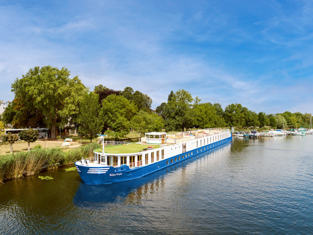 Flusskreuzfahrtschiff mit blauem Rumpf liegt an einem baumbestandenen Flussufer, im Hintergrund ein kleiner Sportboothafen.