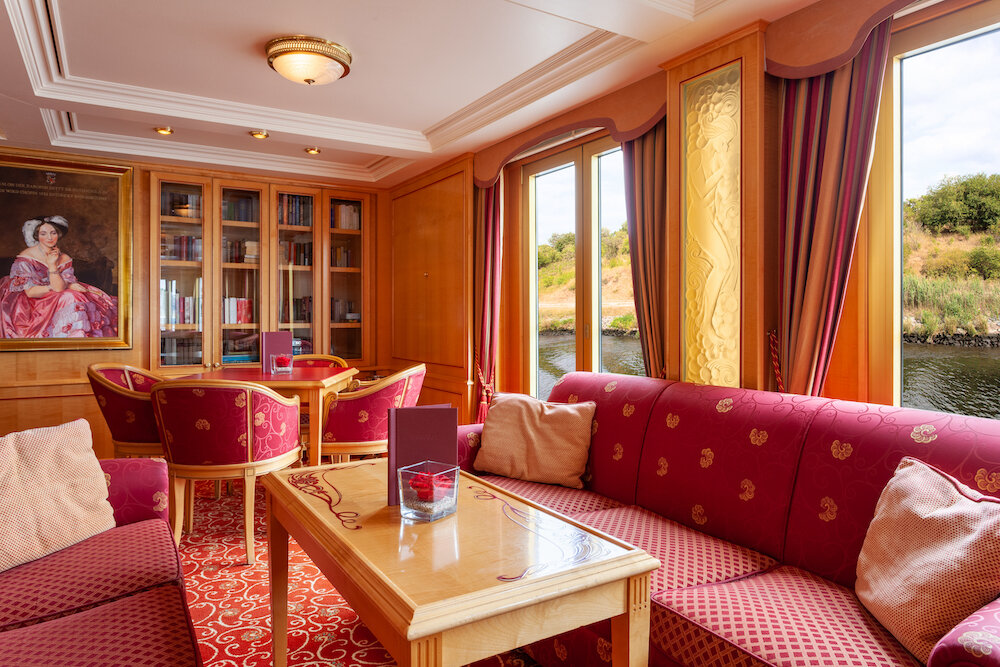 Salon eines Flusskreuzfahrtschiffs mit gepolsterten Sofas, Sesseln, Bücherschrank und Fenstern mit Blick auf das Ufer.