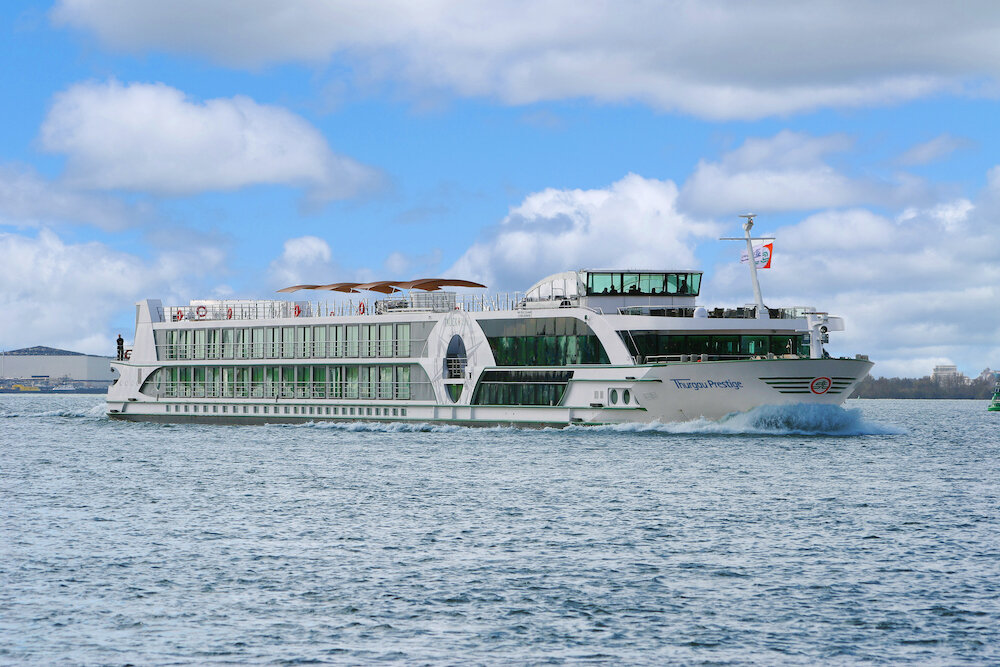 Das Flusskreuzfahrtschiff Thurgau Prestige von Thurgau Travel fährt auf einem Fluss, umgeben von blauem Himmel mit weißen Wolken. Das weiße Schiff mit Panoramakabinen und Sonnensegeln zeigt stolz die Schweizer Flagge am Heck.
