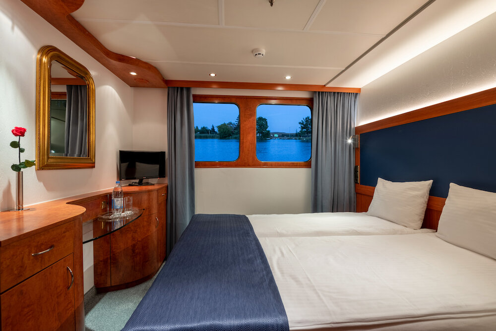 Außenkabine auf dem Hauptdeck eines Flusskreuzfahrtschiffs mit Doppelbett, Holzmöbeln, Spiegel und Fenster zum Flussufer.