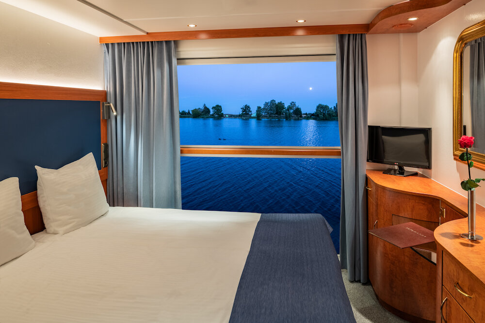 Außenkabine auf dem Oberdeck eines Flusskreuzfahrtschiffs mit Doppelbett und geöffnetem Panoramafenster mit Blick auf den Fluss.