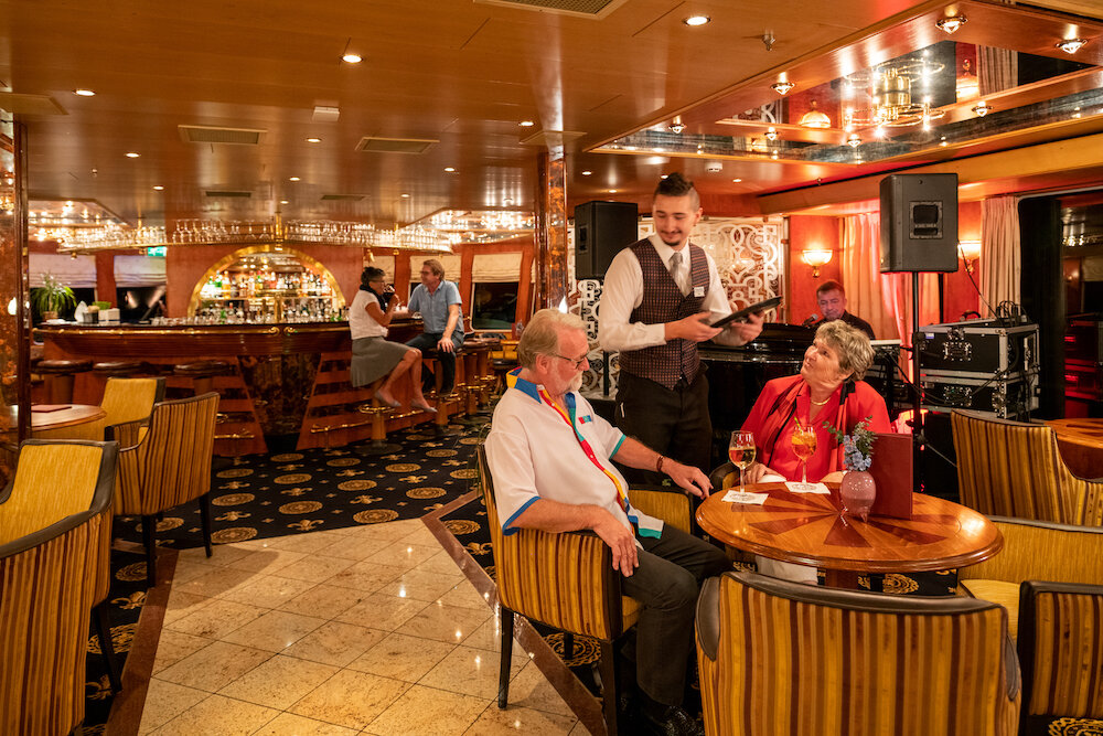 Panorama-Salon eines Flusskreuzfahrtschiffs mit Sitzgruppen, Bartresen im Hintergrund und einem Kellner, der Gäste bedient.