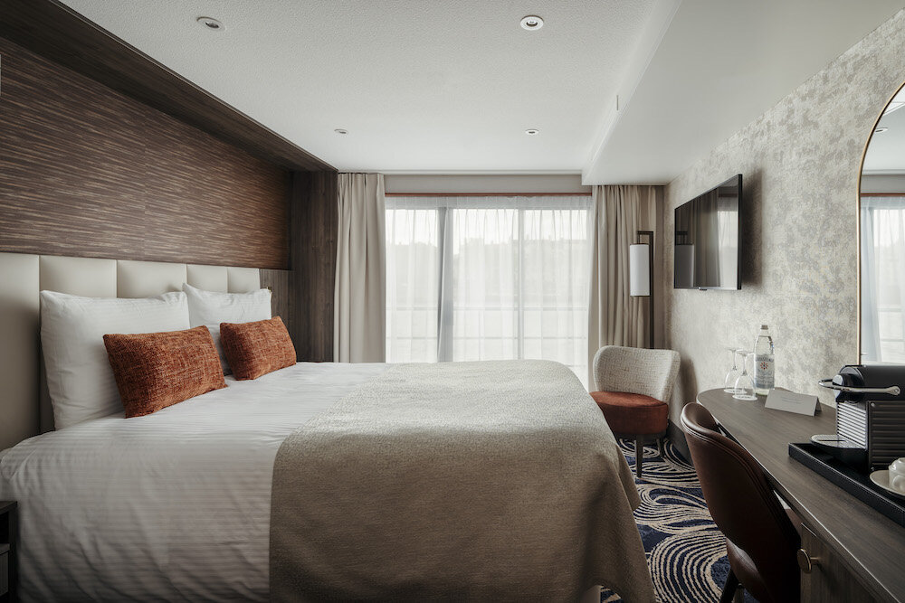 Elegante Doppelkabine auf dem Flusskreuzfahrtschiff Voyage von Thurgau Travel mit großem Doppelbett, weißen Laken, orangen Akzentkissen, beigem Überwurf, dunkler Holzvertäfelung, gepolstertem Kopfteil, bodentiefen Fenstern mit Vorhängen, Wandfernseher, Schreibtisch mit Kaffeemaschine, kleinem Sessel, rundem Wandspiegel und gemustertem Teppich.