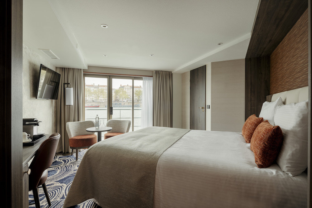 Moderne Junior Suite auf dem Flusskreuzfahrtschiff Thurgau Travel Voyage mit Doppelbett, orangen Dekokissen, Wandfernseher, Sitzecke mit zwei Sesseln und Blick durch bodentiefe Fenster auf die Baseler Wasserfront.