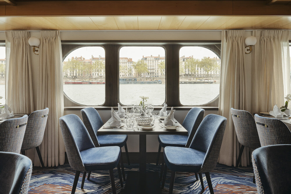 Eingedeckter Esstisch im Restaurant des Flusskreuzfahrtschiffs Voyage von Thurgau Travel mit blauen Samtstühlen, weißen Stoffservietten, Weingläsern und großen Panoramafenstern mit Blick auf einen Fluss und die Skyline von Lyon.