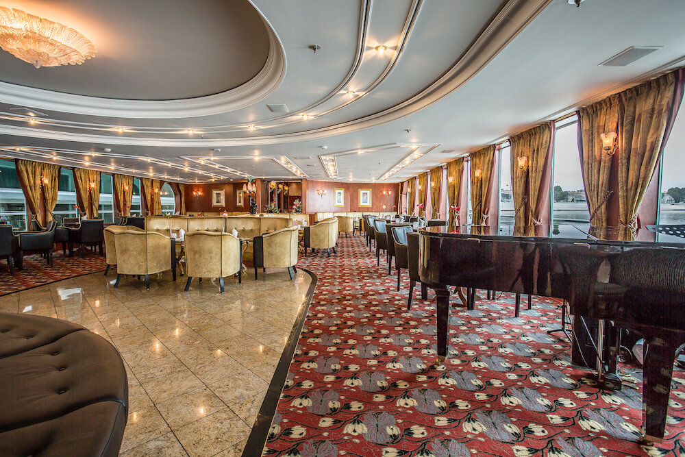 Elegant eingerichteter Salon auf dem Flusskreuzfahrtschiff Thurgau Travel Voyage mit schwarzem Flügel, gemütlichen cremefarbenen Sesseln, rotem gemustertem Blumenteppich, poliertem Marmorboden, goldfarbenen Vorhängen und funkelnden Kristallleuchtern.