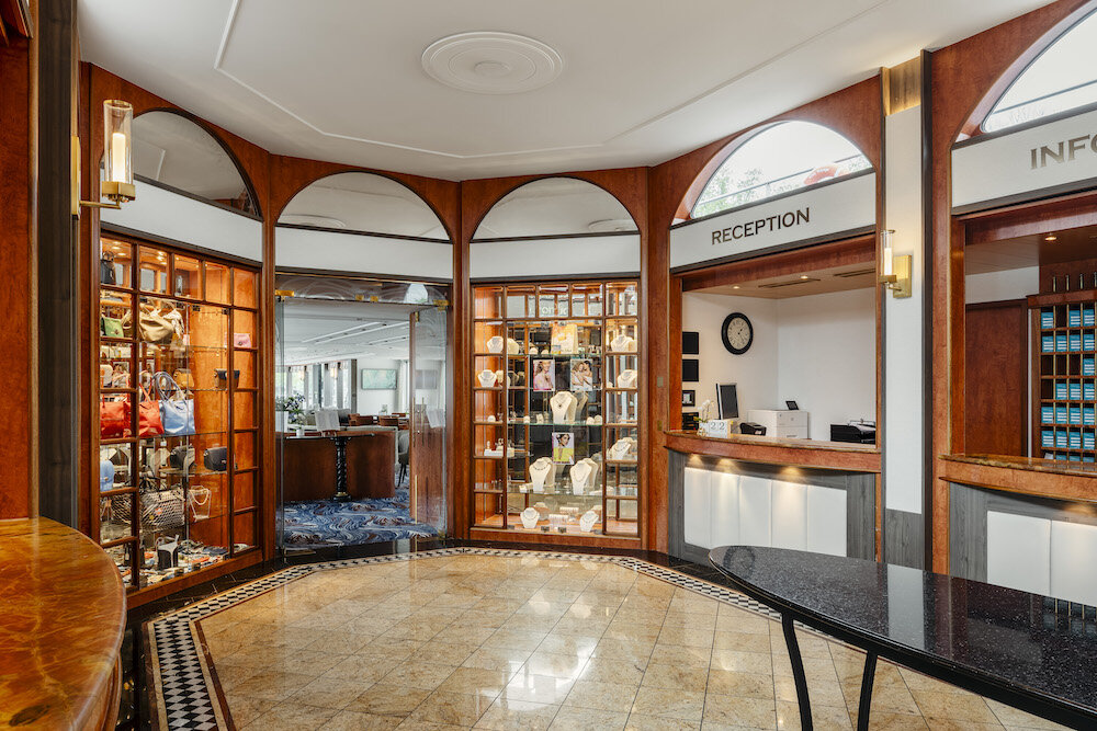 Eingangshalle und Rezeption der MS Voyage von Thurgau Travel mit holzvertäfelten Wänden, verglasten Schmuck- und Accessoire-Vitrinen unter Rundbögen, einer Empfangstheke mit Uhr und Info-Counter sowie poliertem Marmorboden und einem runden Marmortisch im Vordergrund.