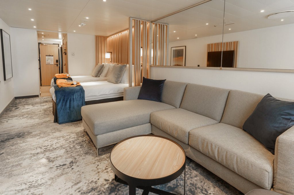 Explorer Suite der Ultramarine mit offenem Wohnbereich, Sofa, Bett hinter Holzlamellentrennwand und Expeditionsausrüstung.