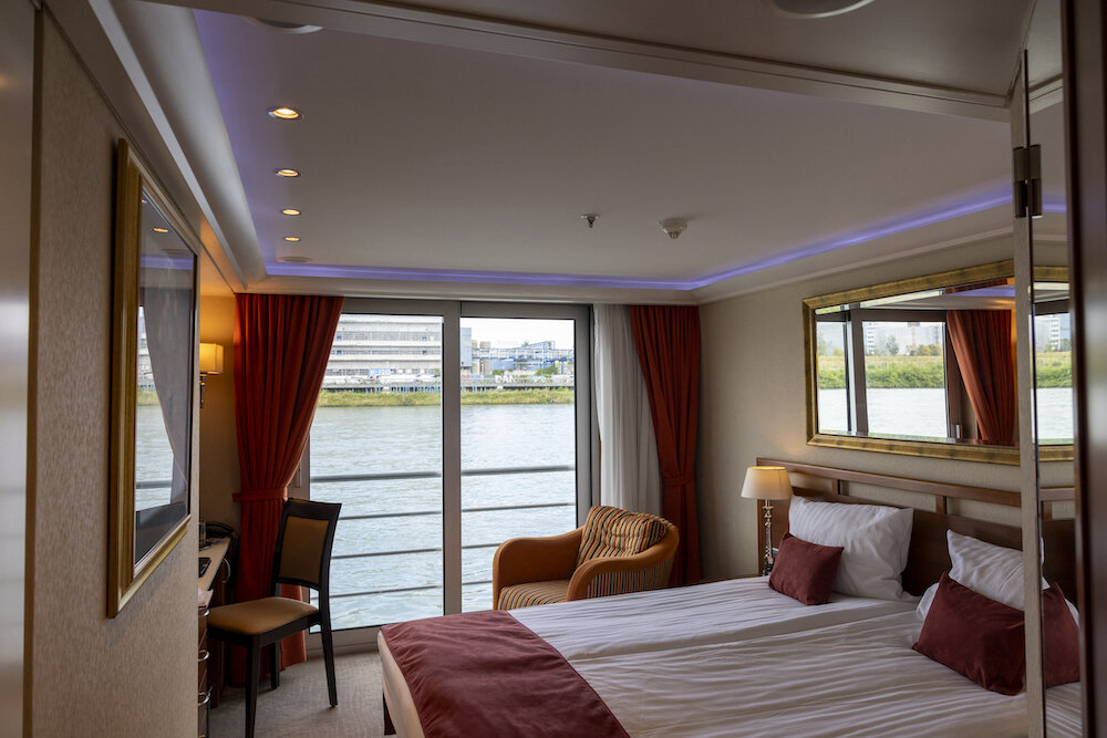 Deluxe-Doppelkabine auf dem Oberdeck mit Doppelbett, Sesselecke und französischem Balkon mit Blick auf den Fluss