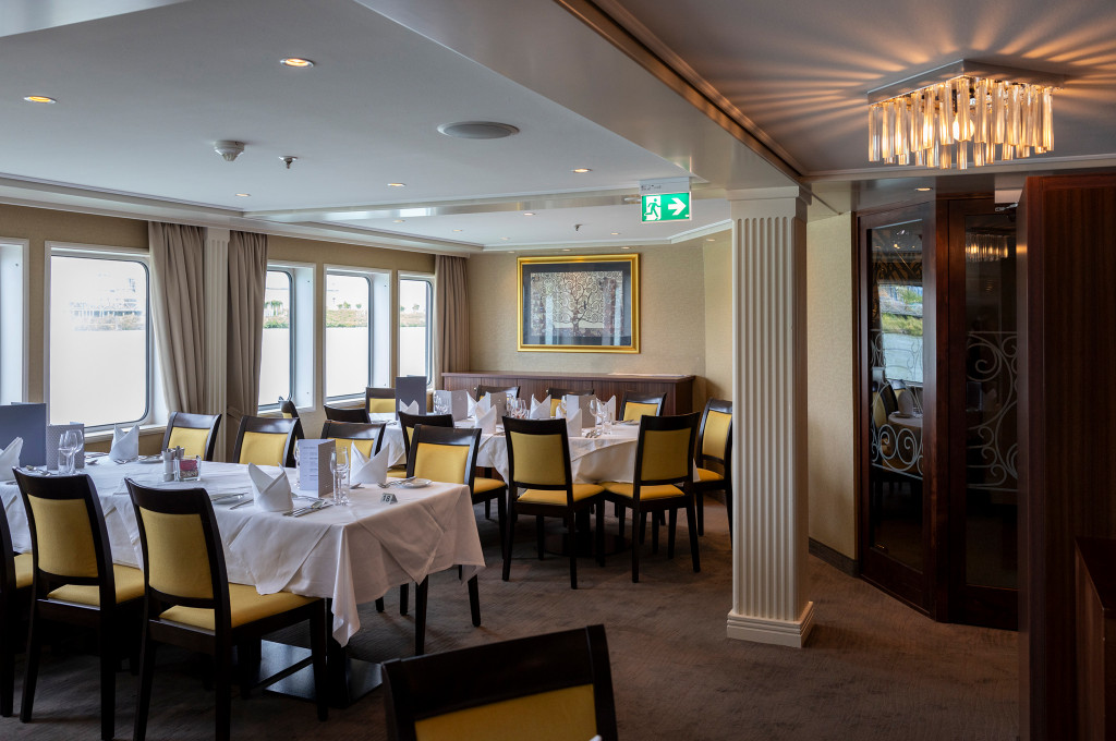 Bordrestaurant mit weiß eingedeckten Tischen, gelben Stuhlpolstern, Kronleuchter und Fensterreihe zum Fluss
