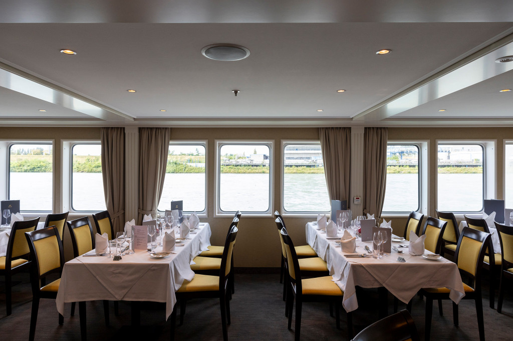 Bordrestaurant mit eingedeckten Tischen, gelben Stuhlpolstern und großer Fensterreihe mit Blick auf den Fluss