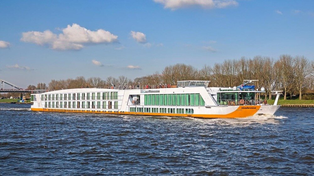 Flusskreuzfahrtschiff in Fahrt auf einem Fluss, seitlich aufgenommen vor einer bewaldeten Uferlandschaft.