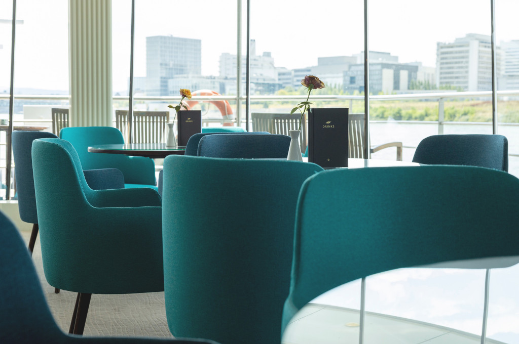 Lounge mit petrolfarbenen Sesseln und kleinen Tischen vor bodentiefen Panoramafenstern mit Blick auf den Fluss