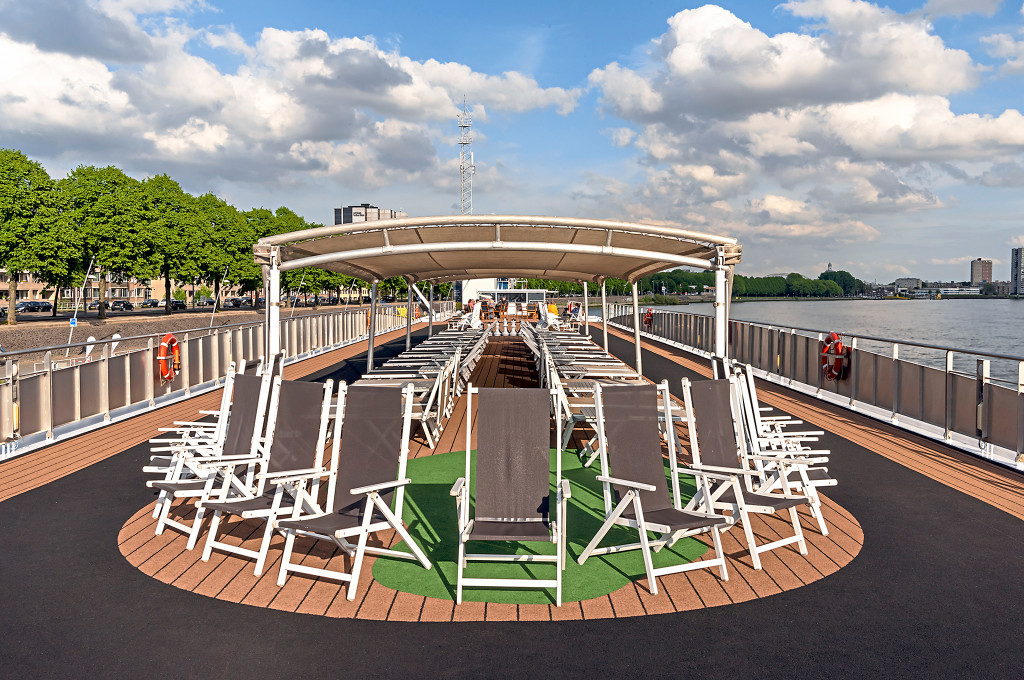 Sonnendeck eines Flusskreuzfahrtschiffes mit Liegestühlen, Kunstrasenfläche und überdachtem Bereich am Flussufer