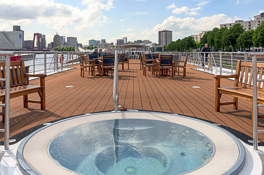 Sonnendeck eines Flusskreuzfahrtschiffes mit Whirlpool im Vordergrund, Holzbänken und Sitzgruppen vor der Stadtkulisse