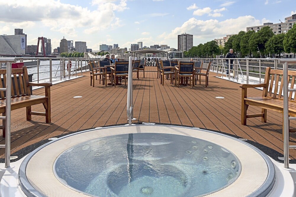 Sonnendeck eines Flusskreuzfahrtschiffs mit Whirlpool, Holzstühlen und Sitzbänken vor der Skyline von Rotterdam.