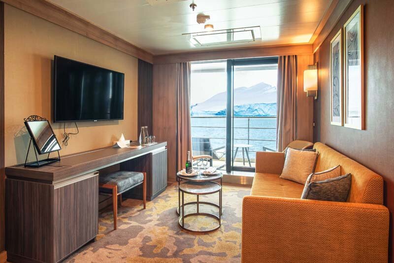 Deluxe Suite auf der World Explorer mit Sofa, Fernseher und Balkon mit Blick auf eine arktische Gletscherlandschaft.