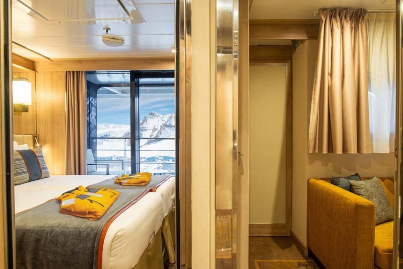 Triple Suite auf der World Explorer mit zwei Betten, gelbem Expeditionsparka und arktischer Bergkulisse vor dem Fenster.