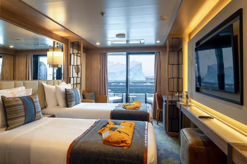 Veranda Suite auf der World Explorer mit zwei Betten, Expeditionsparkas und Blick auf Eisberge durch die Verandatür.