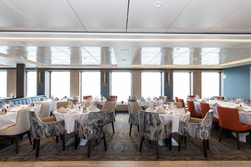 Restaurant auf der World Explorer mit gedeckten Tischen, gemusterten Polstersesseln und Panoramafenstern.