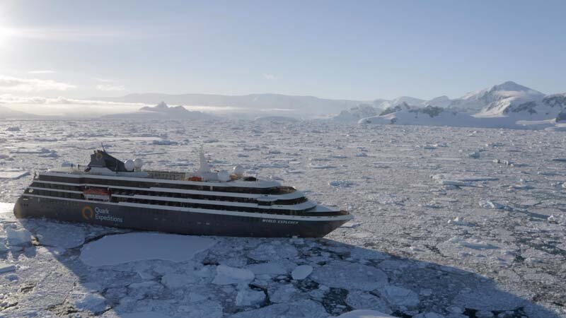 Expeditionsschiff navigiert durch Packeis in der Antarktis mit schneebedeckten Bergen im Hintergrund