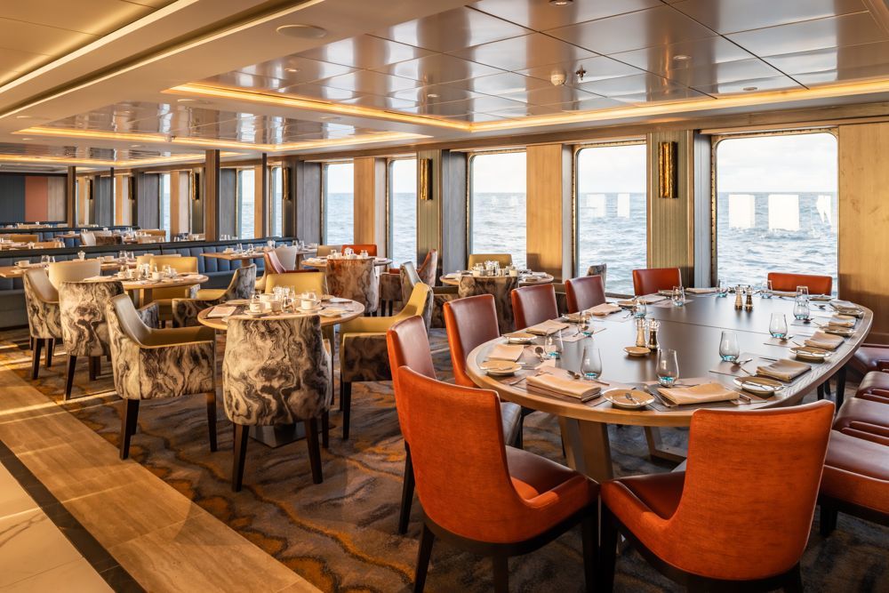 Modernes Restaurant mit runden Tischen, gemusterten Sesseln und großen Fenstern zum Meer auf einem Expeditionsschiff